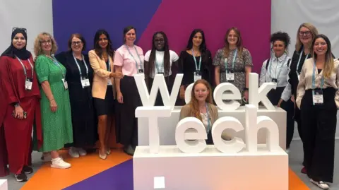 Eleven smiling colourfully dressed women, wearing lanyards are posed behind large white 3D 'Tech Week' signage with another woman seated between the words 'Tech' in the foreground, and 'Week', in front of the row of smiling women.