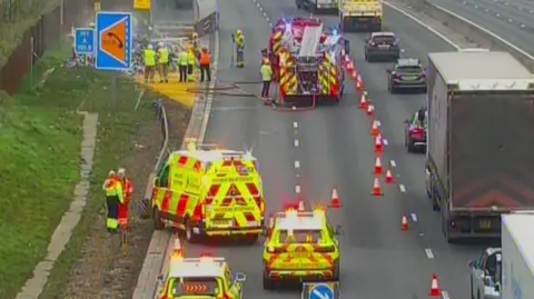 Two lanes of traffic have been blocked by a police car. A lorry has been pulled to the side of the road and smoke is filling the air behind it. a fire engine and water tanker are at the scene.