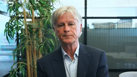 James, a man in a dark suit jacket and light shirt sits in an office space with tall buildings visible through the windows and a plant behind him. He has white hair and looks at the camera.