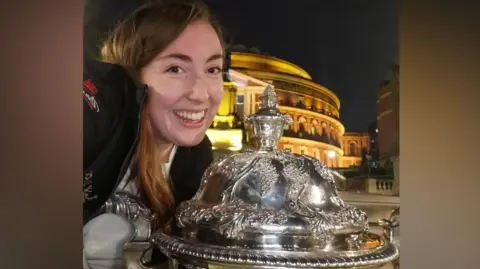 Stephanie Binns Stephanie Binns poses with a trophy outside the Royal Albert Hall. She is smiling. It is night time.