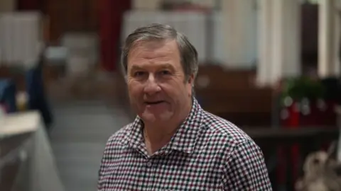 Jamie Niblock/BBC Phil Judd stands in a church. He wears a checked shirt and is looking at the camera lens. 