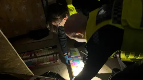 Two PCSOs in uniform leaning down with flashlights in a dark room where illegal vape products in colourful boxes are lined up below them on the floor