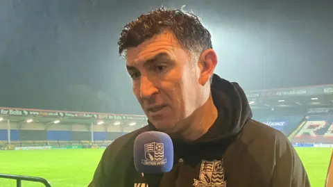 Warren Muggleton/BBC Southend United head coach Kevin Maher being interviewed after the game has been abandoned. His hair is wet and he is speaking into a microphone. Behind him there is heavy rainfall lashing down on the pitch and empty stands.