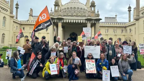 A group of workers hold banners and signs outside an Indo-Islamic-style building. 
