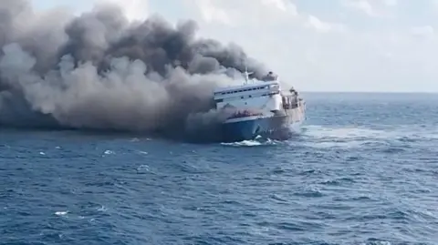 A still shot of the KMP Mutiara Sentosa with smoke billowing from behind as the passenger vessel slants to the side in the ocean