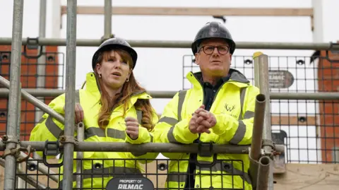 PA Media Sir Keir Starmer and Angela Rayner wearing hard hats and hi-visability jackets as they lean on scaffolding, looking out across a construction site for a housing development