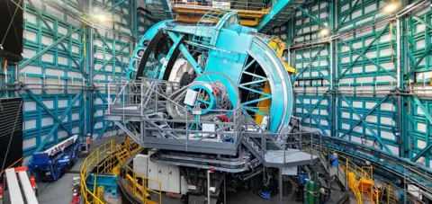 RubinObs A round blue ring with cables coming off it and a metal platform leading up to it. It is inside a domed building with blue and white railings around it. The blue ring is elevated on a platform with metal stairs leading up to it. Around it there is construction equipment.