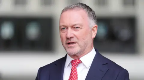 PA A man wearing a navy suit, white shirt and red tie is looking at a camera.