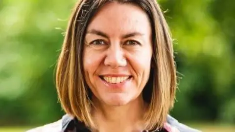 Sadie Parker, director of primary care for NHS Norfolk and Waveney, looks straight at the camera. She has a long dark blonde bob and is smiling. Her black shirt is decorated with flowers