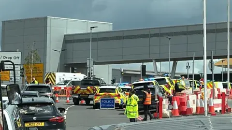 A queue of cars runs to the side of a police sign and emergency vehicles and workers.