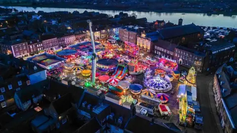 King's Lynn Mart Fair A view from a drone of King's Lynn's town centre with the Mart set up on the Tuesday Market Place. Its rides are lit up in a rainbow of colours. Behind the town is the river Great Ouse and a beautiful orange sky. 