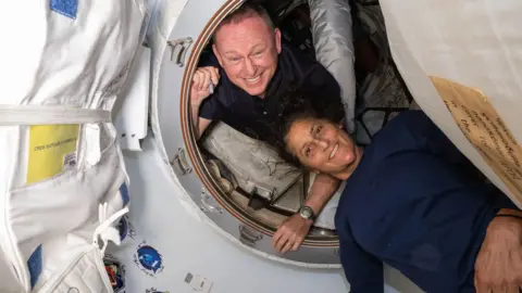 Os astronautas da NASA Butch Wilmore e Suni Williams a bordo da estação espacial internacional sorrindo enquanto posam no laboratório em órbita. Butch está cuidando de uma escotilha circular branca, enquanto Suni está deitado do lado de fora dele. 