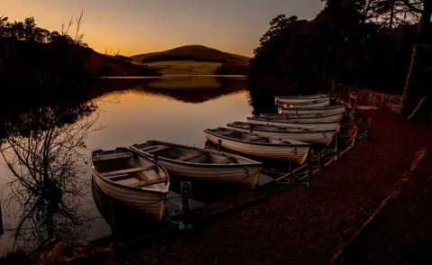 Stewart Roberts Seven small boats lined up on the bank of a reservoir during sunset