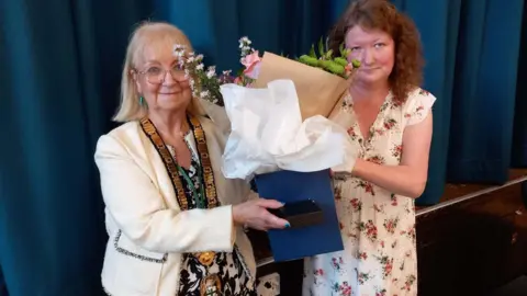 BBC Councillor Sue Eacock, left, pictured with Councillor Joanna Kane when she became the new Mayor of Redditch - Councillor Eacock is wearing a white jacket with a large gold chain around her neck. She is being handed flowers to mark the occasion.