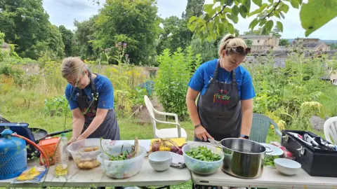 Bath Mind Two people cooking on two tables covered in foraged food. They are in a green garden on what seems like a sunny day.