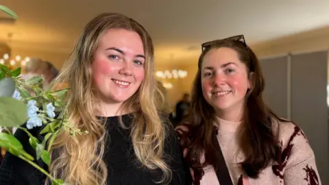 Chloe and Emma Hayden are looking straight ahead and both are smiling. Chloe on the left has long blonde hair and a dark crew neck jumper. Emma, Chloe's mother has long dark hair and has sunglasses on the top of her head. She is wearing a light pink jumper with a purple patteren.
