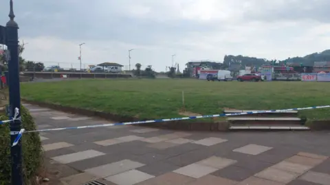 Police tape covers an pathway with a grass area behind and some vehicles.