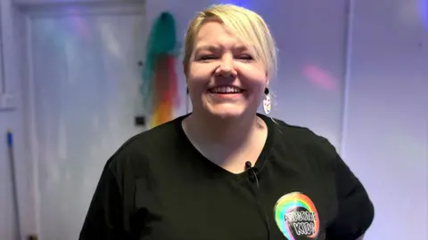 BBC Pamela Baxter has short blonde hair and is wearing a black T-shirt. On the left hand side there is a logo with a rainbow swirl and black writing reading "AUsome Kids"