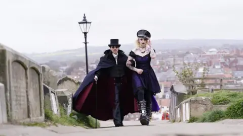 Danny Lawson/PA A man and a woman dressed in gothic clothing walk arm in arm through the graveyard near Whitby Abbey