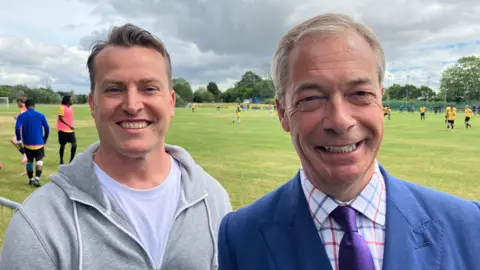 James McMurdock and Nigel Farage smile and look directly into the camera as they stand by the side of a football match. 