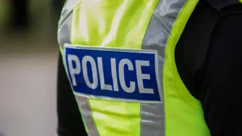 A close-up of someone's back. They are wearing a black long-sleeved top under a yellow hi-viz vest with grey stripes marked "POLICE".