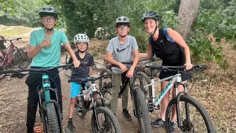 Leanne Brown/BBC A family of four standing on bikes with helmets on in front of greenery and a dirt track, looking into the camera. An 11-year-old boy wearing a turquoise T-shirt and glasses holds a thumbs-up. To his right, a seven-year-old boy wearing a dark grey T-shirt and bright blue shorts smiles. To his right, another 11-year-old boy wearing glasses and a light grey T-shirt with a Nike tick on it smiles, next to his mum who is wearing a black tank top and shorts, with her hand on his back and smiling.