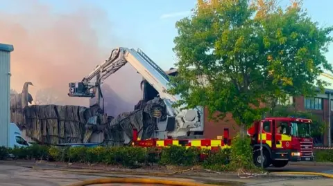 A burnt down building, with an aerial ladder appliance outside, a tree to the right and a building behind it. Smoke is rising in the air. 