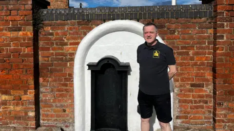 David Purchase standing in front of the drinking fountain. He is wearing a balck t-shirt and shorts, with his arms posed behind his back. Behind him there is a white painted arch on a red brick wall. Within the arch there is a cast iron back-plate with an inscription on the trough at street level, that reads Gloucester Local Board Of Health 1863.