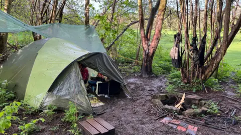 A green tent and tarpaulin tied between trees in a small woodland beside an open green space. There is a campfire area marked out by stones.