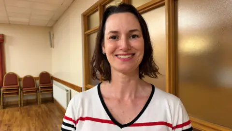 Daniella Mediondo de Lima has medium length dark brown hair and is wearing a white sweather with one red and several navy stripes and a navy-trimmed V neck. She is standing in a hall with a glass partition wall behind her.