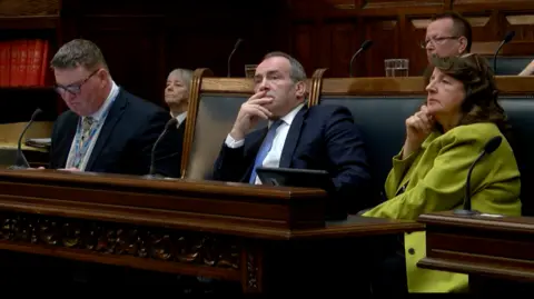 Chief Minister Alfred Cannan has short greying hair and is sitting in a dark suit, white shirt and blue tie, in Tynwald, surrounded either side by his political colleagues. The large seats are oak with dark green leather cushioning, which are lined with an ornate oak table in front.