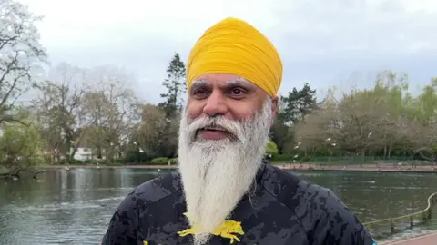 Manny Singh Kang - a man with a long grey beard and yellow turban stands next to a lake in West Park, Wolverhampton