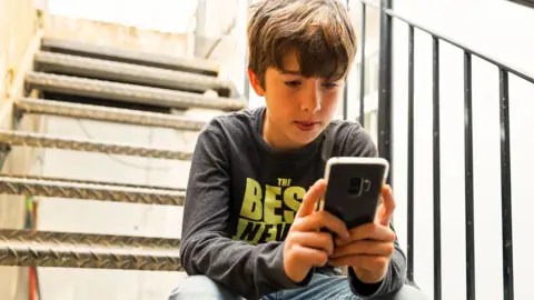 Boy aged about 14 looking at his phone. He is sitting on metal steps outside and wearing a grey t-shirt with neon yellow writing. 