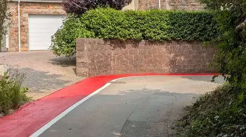 Andy Jay The picture shows a tight bend in a road with a bright red painted pedestrian walk way. The path has a white line to mark the pedestrian zone. There is a house and a driveway in the background with plenty of greenery.
