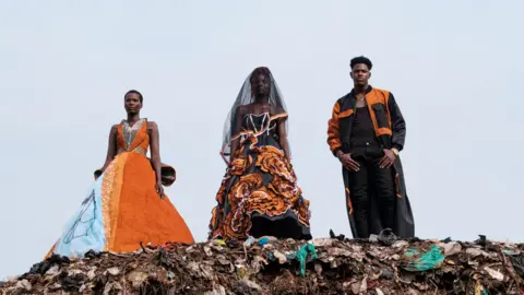 KHABIR DHANJI/AFP Models wearing black and orange clothes stand on top of a rubbish heap in Kampala, Uganda - Saturday 20 July 2024