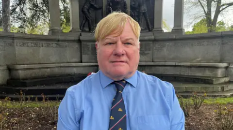 Neil Atkinson, a man with blond hair covering his forehead. He is stood in front of the memorial and is wearing a blue shirt with a navy blue and red tie.