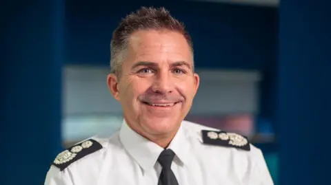 A Simon Tuhill pictured smiling with short dark spikey hair. He is wearing a white shirt and black tie with black Epaulettes on his shoulders.