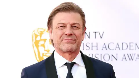 PA Media Close-up image of Sean Bean looking into the camera, wearing a dark blue and black tuxedo and black tie.