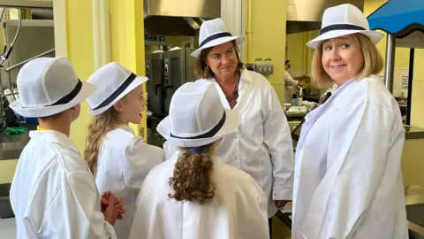 BBC A smiling Eluned Morgan, Lynne Neagle and three pupils all wearing white coats in front of a school kitchen