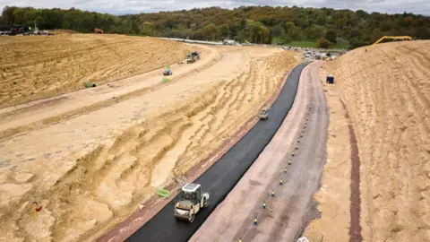 National Highways Rollers squashing Tarmac on a new road being built between two sections of rock and soil which have been made in to large mounds. 