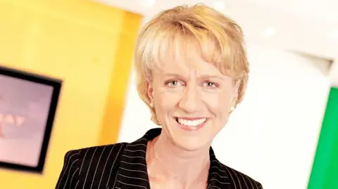 A smiling Sally Taylor wearing a black pinstripe suit. She has short blonde hair and grey eyes.