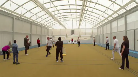 A large sports hall with white walls, an arched roof and yellow floor with people standing around and cricket stumps in the foreground