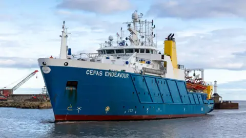 The research vessel Cefas Endeavour coming in to harbour. It has a blue hull and white wheelhouse. On the wheelhouse roof are many piece of radar and satellite technology. Lifeboats can be seen towards the stern. The sea is calm and there is a quay behind the boat.