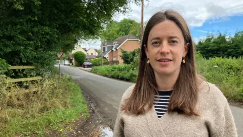 Amy Holmes/BBC A woman with long brown hair and a being cardigan is standing by a quiet country road, with houses, trees, and a give-way sign in the background under a partly cloudy sky.