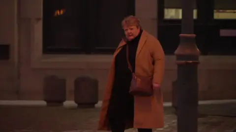 A woman arrives at court wearing a long orange coat. She was short blonde hair