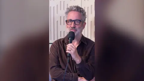 David Baddiel sat wearing a brown shirt holding a black mic.