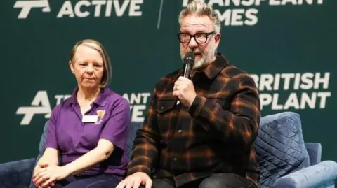 British Transplant Games Woman in a purple t-shirt sitting on a blue sofa next to Sheffield artist Pete McKee, in a brown tartan shirt and black glasses holding a microphone, with the British Transplant Games banner behind