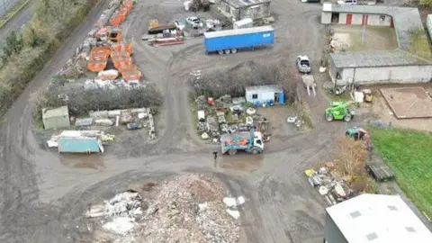 Environment Agency Drone image of a farm yard with a large pile of rubble, and areas of skips, appliances and assorted waste, separated by muddy roads and a number of vehicles