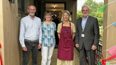 Wiltshire Council Four people stood next to each other and smiling at the camera. There are two women in the centre and a man on each end of the line. The woman on the right is wearing a burgundy apron and a dress and the other woman is wearing white trousers and a blue patterned top. The man on the right is wearing a suit and the other man is wearing a shirt and dark trousers. 