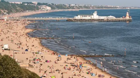 Pier stretching out to see with sandy beach packed with beachgoers curving round to the right.